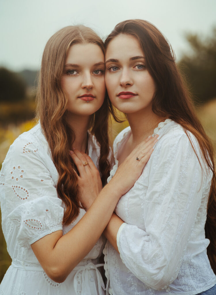 Dwie młode kobiety w białych sukienkach stojące blisko siebie na tle natury, patrzące wprost w obiektyw — artystyczna fotografia portretowa w plenerze