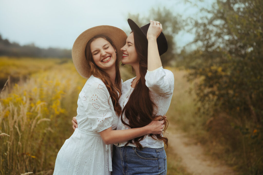 Dwie młode kobiety w białych ubraniach i kapeluszach obejmujące się z uśmiechem na polnej ścieżce — naturalna fotografia portretowa w plenerze
