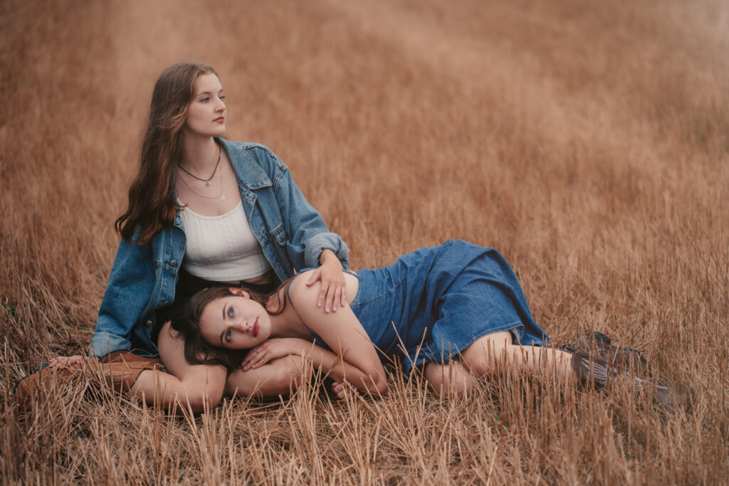 Dwie młode kobiety w jeansowych stylizacjach odpoczywające na polu po żniwach, jedna leży z głową na kolanach drugiej — artystyczna fotografia portretowa w plenerze.