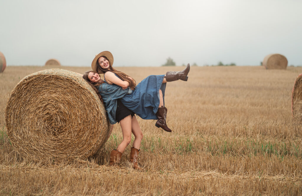 Dwie młode kobiety w stylizacjach boho i kowbojskich butach bawiące się na polu ze słomianymi belami — radosna fotografia portretowa w plenerze.