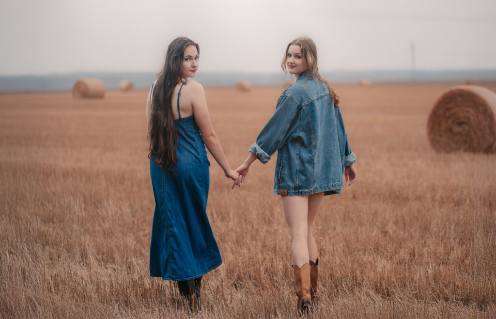 Dwie młode kobiety trzymające się za ręce na polu ze słomianymi belami, w jeansowych stylizacjach boho — naturalna fotografia portretowa w plenerze