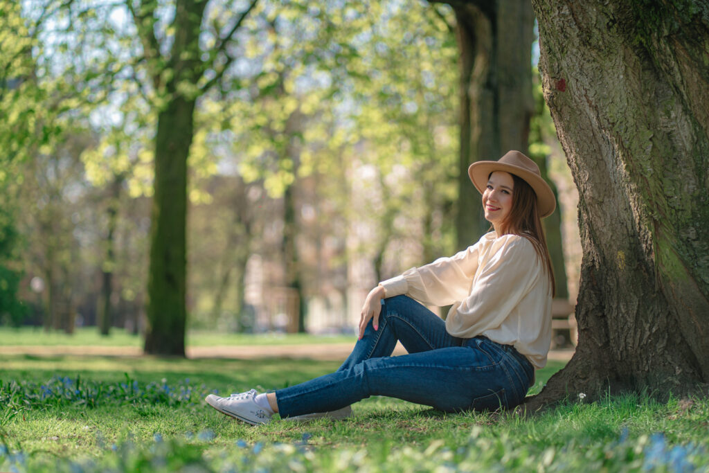Kobieta w kapeluszu, jasnej bluzce i dżinsach siedząca pod drzewem w parku w słoneczny dzień — naturalna fotografia portretowa w plenerze