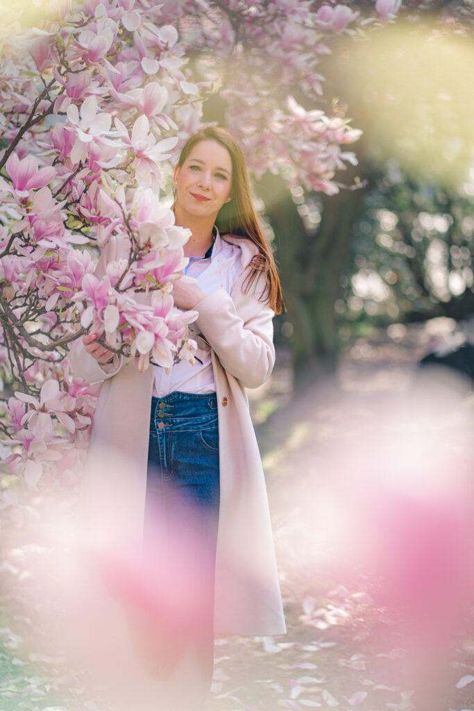 Kobieta w jasnym płaszczu i dżinsach stojąca w ogrodzie pełnym kwitnących magnolii, romantyczna fotografia portretowa w plenerze