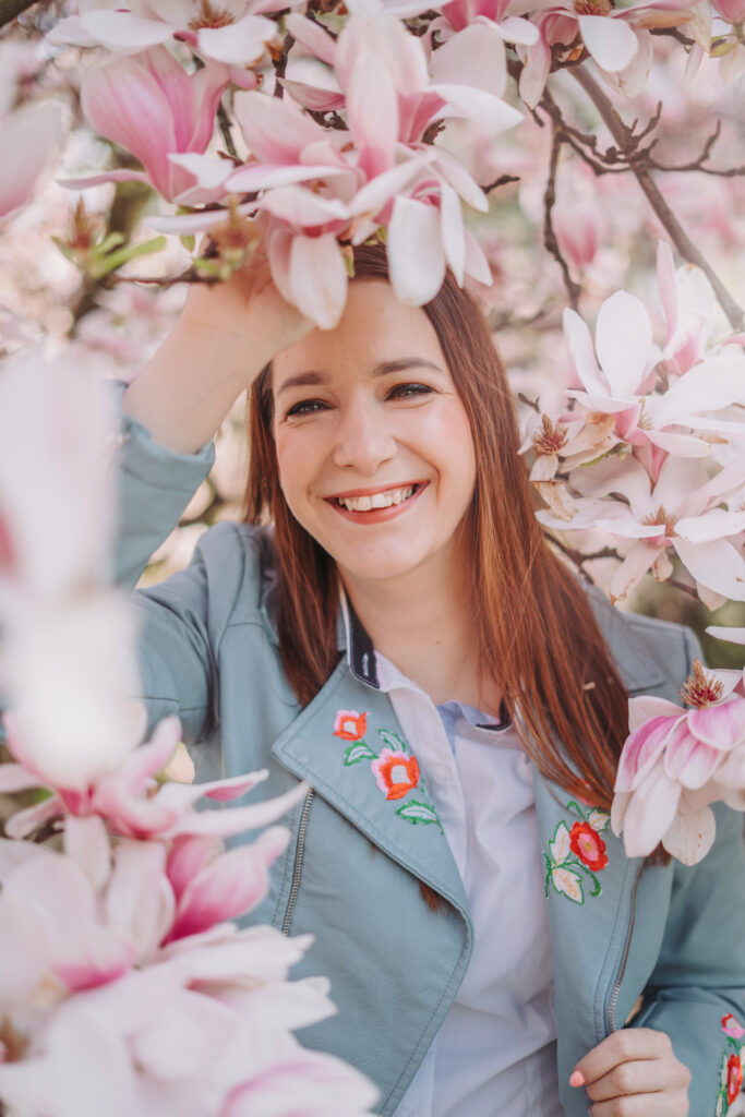 Uśmiechnięta kobieta w turkusowej kurtce z haftem kwiatowym, stojąca wśród kwitnących magnolii — wiosenna fotografia portretowa w plenerze.