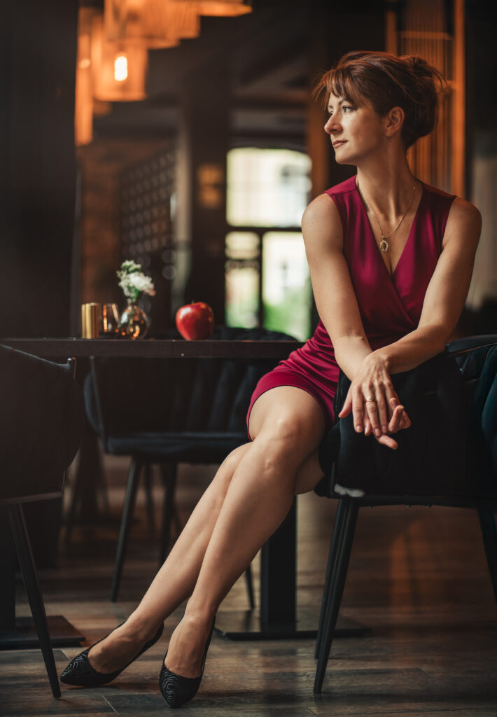 Kobieta w eleganckiej czerwonej sukience siedząca w stylowej kawiarni, oparta o krzesło i patrząca w bok — profesjonalna fotografia portretowa w klimatycznym wnętrzu.