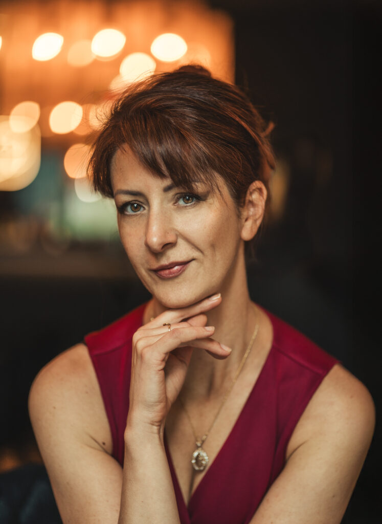 Kobieta w czerwonej sukience na portrecie w kawiarni, z delikatnym uśmiechem i dłonią opartą o brodę — profesjonalna fotografia portretowa w klimatycznym wnętrzu.