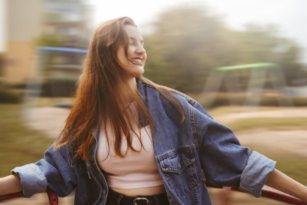 Uśmiechnięta młoda kobieta w dżinsowej kurtce i białym topie, kręcąca się na karuzeli w plenerze — dynamiczna fotografia portretowa w naturalnym świetle.