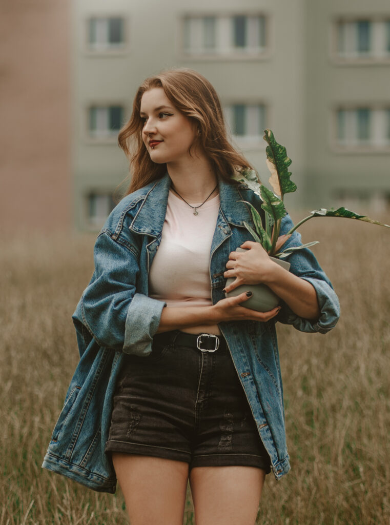 Młoda kobieta w dżinsowej kurtce i czarnych szortach trzymająca doniczkę z rośliną, stojąca na tle wysokiej trawy i budynków — naturalna fotografia portretowa w plenerze.