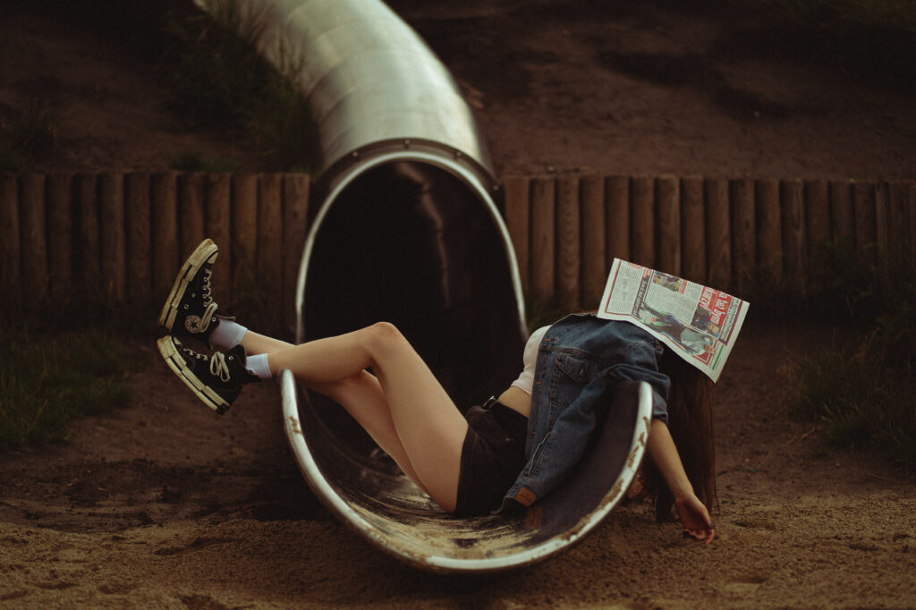 Młoda kobieta w trampkach i dżinsowej kurtce leżąca na metalowej zjeżdżalni z gazetą na twarzy — kreatywna fotografia portretowa w plenerze