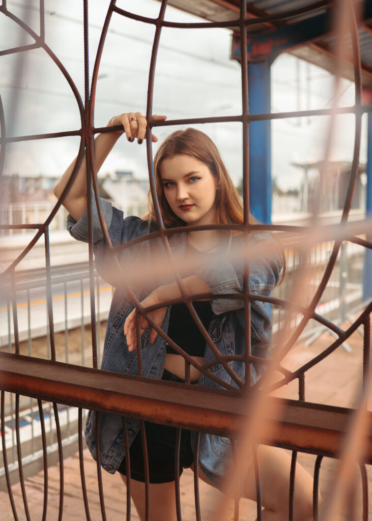 Młoda kobieta w dżinsowej kurtce i czarnym topie, pozująca przy metalowej konstrukcji na tle dworca kolejowego — nowoczesna fotografia portretowa w przestrzeni miejskiej.