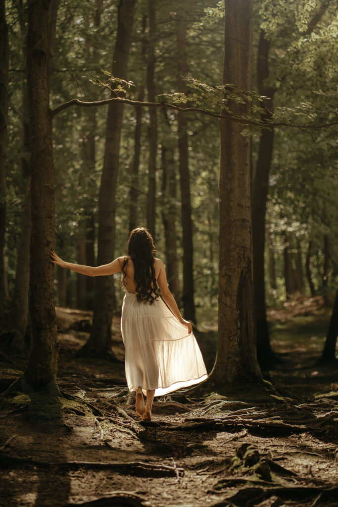 Kobieta w białej, zwiewnej sukience idąca boso przez las, fotografia portretowa w naturalnym świetle — profesjonalna sesja zdjęciowa w plenerze