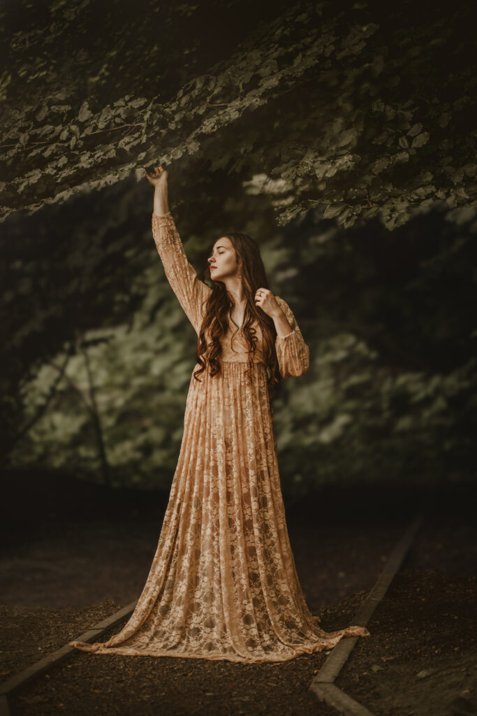 Kobieta w długiej, koronkowej sukni w odcieniu beżu, stojąca w lesie i unosząca rękę ku gałęziom drzewa — artystyczna fotografia portretowa w plenerze