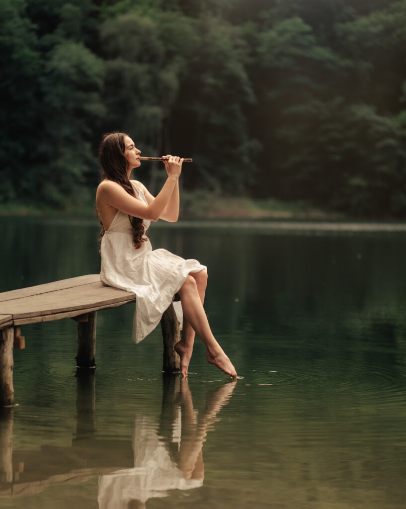 Kobieta w białej sukience siedząca na drewnianym pomoście nad jeziorem, grająca na flecie — artystyczna fotografia portretowa w plenerze.