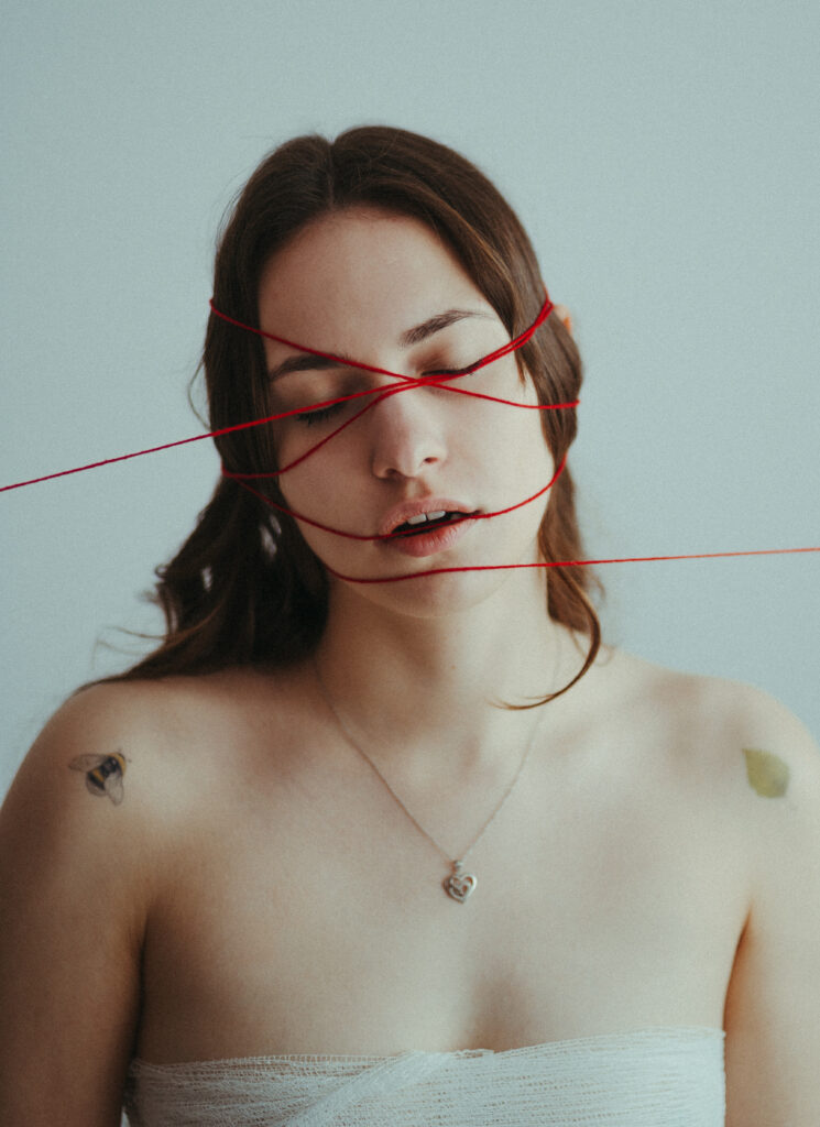 portret artystyczny kobiety fotografia konceptualna czerwona nić na twarzy symbolika nici losu motyw przeznaczenia w sztuce kobieta z zamkniętymi oczami minimalistyczny portret fotografia z przesłaniem artystyczna fotografia kobieca emocjonalny portret