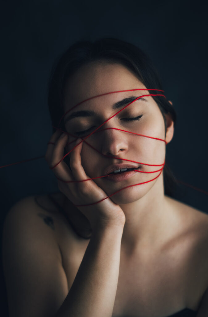 portret artystyczny kobiety fotografia konceptualna czerwona nić na twarzy symbolika nici losu motyw przeznaczenia w sztuce kobieta z zamkniętymi oczami minimalistyczny portret fotografia z przesłaniem artystyczna fotografia kobieca emocjonalny portret