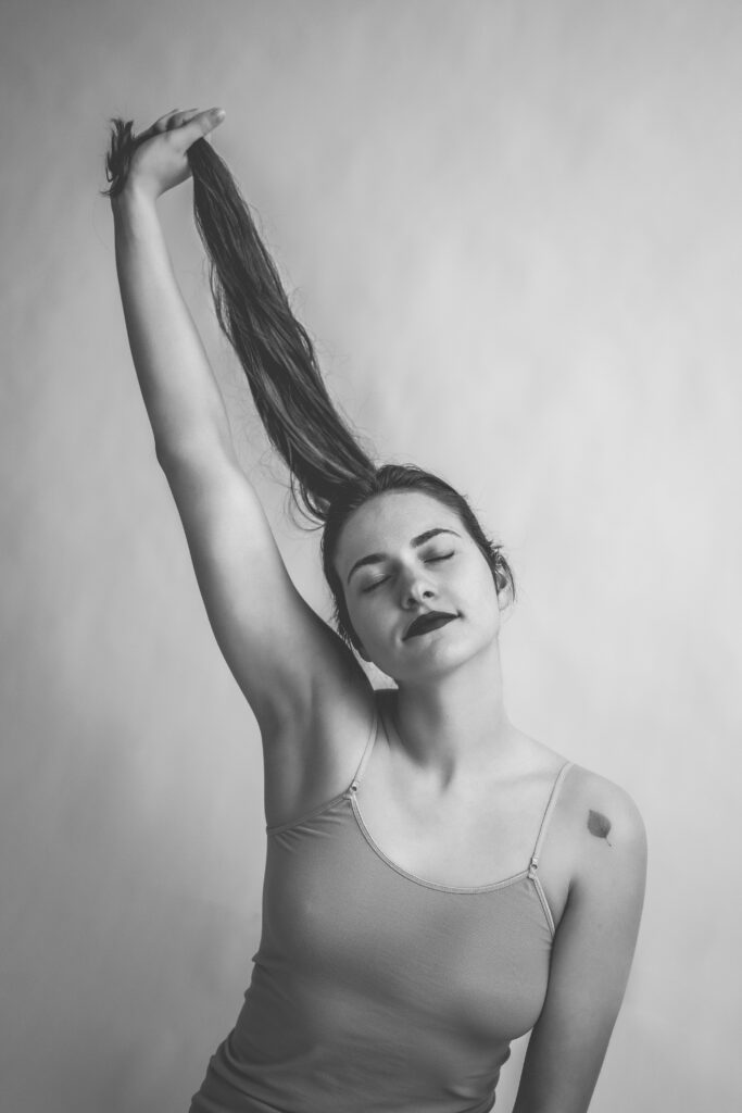 Czarno-biały artystyczny portret kobiety z zamkniętymi oczami, unoszącej rękę i trzymającej włosy, w minimalistycznym, studyjnym ujęciu podkreślającym ekspresję i subtelność